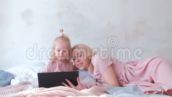 年轻迷人的金发女人用躺在床上的平板电脑教她穿着粉红色裙子的迷人小女儿视频的预览图