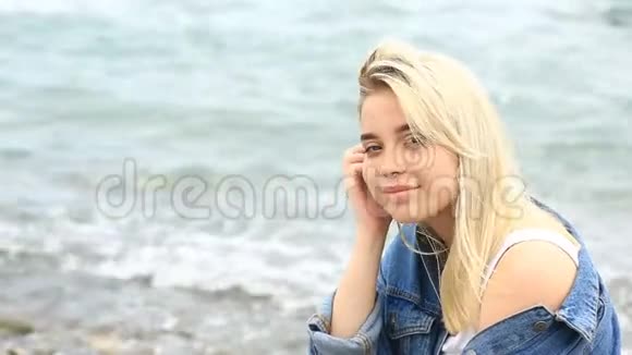 美丽的金发女郎坐在沙滩上可爱的微笑着看着海浪背景下的镜头视频的预览图