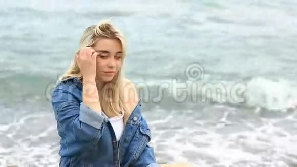 美丽的金发女郎坐在沙滩上可爱的微笑着看着海浪背景下的镜头视频的预览图