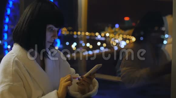 一个穿着白色长袍的年轻漂亮的女人坐在窗边看着手机写着短信从视频的预览图