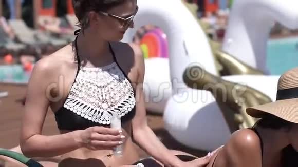 穿比基尼的女孩在她朋友的背上放防晒霜美丽的女孩晒日光浴在游泳池边玩得开心慢动作视频的预览图