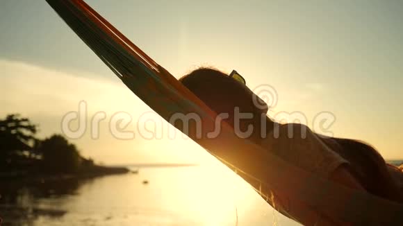 年轻女子在海边海滩的吊床上使用手机背景是美丽的日落戈芬加视频的预览图