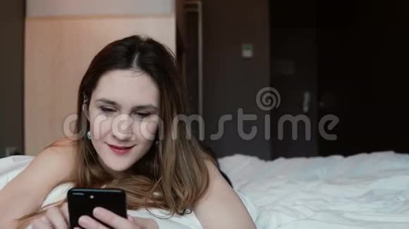 年轻的女人一边用智能手机一边躺在床上微笑女孩触摸屏幕浏览互联网视频的预览图