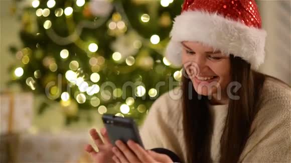 圣诞老人帽子里的女人用小玩意坐在圣诞树旁边长着深色头发的快乐女孩视频的预览图