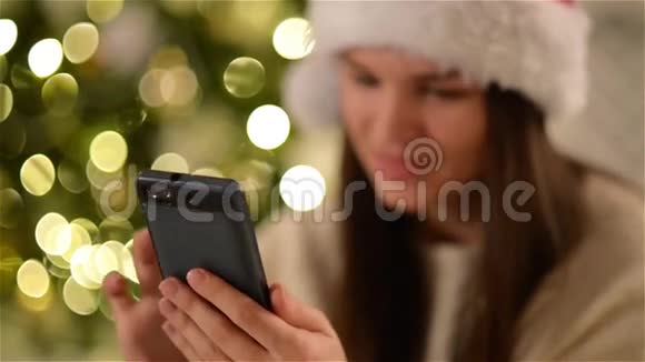 圣诞帽中可爱的布鲁内特在智能手机上发短信在圣诞树背景上微笑长发快乐的女人视频的预览图