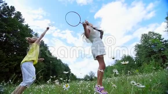 在绿色的洋甘菊草坪上女孩孩子打羽毛球他们跑跳到处乱跑他们玩得很开心夏季视频的预览图