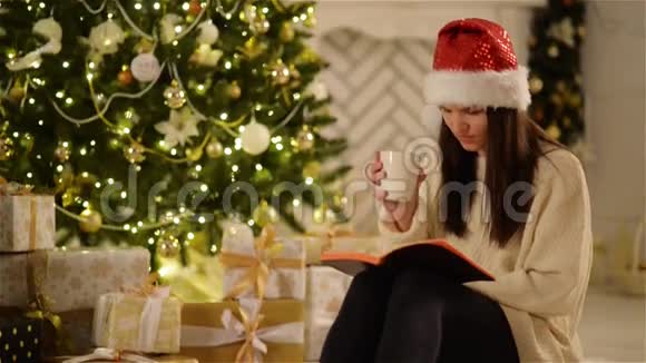 美丽的年轻女子戴着圣诞帽坐在家里一边喝热饮料一边读一本书美丽的布鲁内特视频的预览图