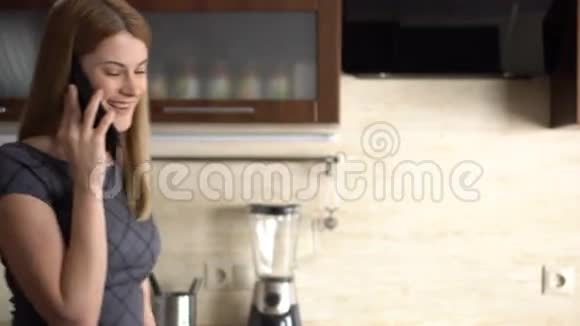 美丽迷人的年轻女子穿着灰色连衣裙站在厨房里用手机说话视频的预览图