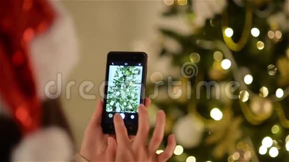 圣诞帽里的女孩手里拿着智能手机拍下了圣诞树的照片美丽的圣诞装饰视频的预览图