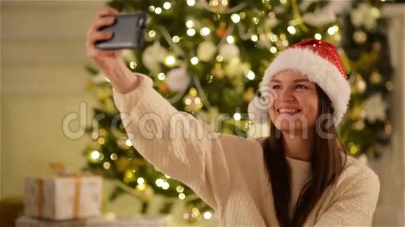 戴着圣诞帽在手机上做自拍的微笑女人的圣诞画像长着深色头发的可爱女士视频的预览图