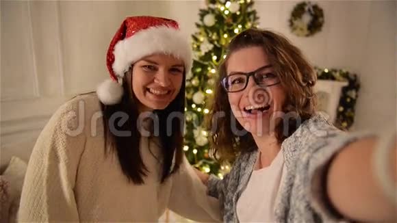 两个微笑迷人的女人在圣诞树背景上做自拍一个长着卷发戴眼镜的女孩视频的预览图
