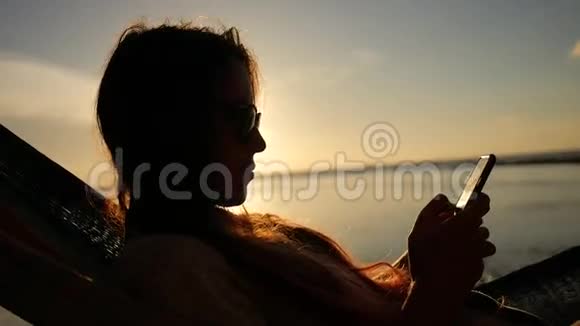 年轻女子在海边海滩的吊床上使用手机背景是美丽的日落戈芬加视频的预览图