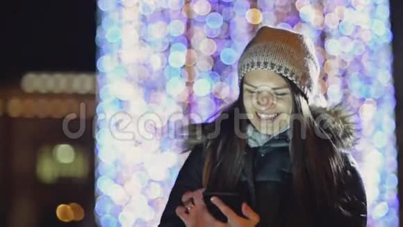 戴帽子的年轻女士站在街上用手机圣诞灯上戴着小玩意的微笑女孩视频的预览图