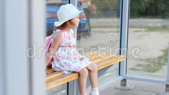 一位戴着帽子背着背包的女游客坐在公共汽车站的长凳上等待交通的进一步发展视频的预览图