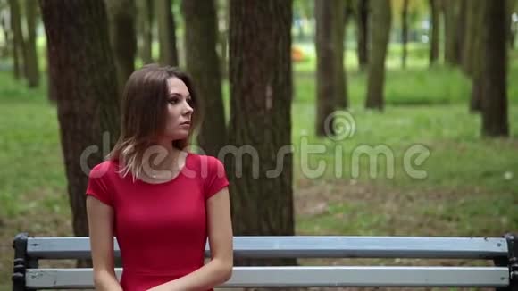 穿着红色裙子的年轻漂亮女孩正坐在公园的长凳上视频的预览图