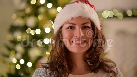 圣诞帽里的卷发女人带着迷人的微笑看着圣诞树背景上的相机特写肖像画视频的预览图