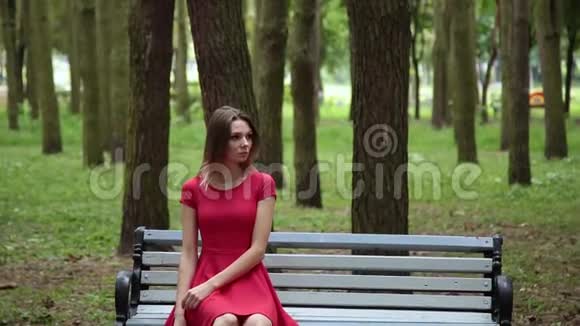 穿着红色裙子的年轻漂亮女孩正坐在公园的长凳上视频的预览图