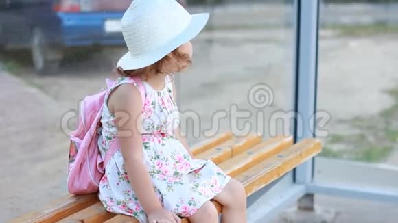 一位戴着帽子背着背包的女游客坐在公共汽车站的长凳上等待交通的进一步发展视频的预览图