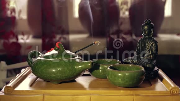 佛像在木茶板上用绿茶壶和杯子燃烧着的香枝散发出的香气视频的预览图