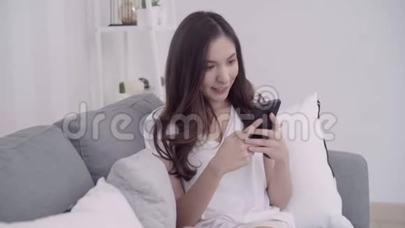 漂亮的亚洲女人在客厅里躺在沙发上用智能手机生活方式女性在家里的概念视频的预览图