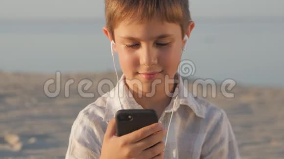 特写男孩在耳机上听音乐10岁的孩子喜欢坐在沙滩上的智能手机视频的预览图