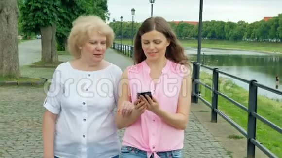 快乐的老年妇女和年轻妇女沿着海滨散步使用智能手机成年母亲和女儿在公园散步视频的预览图