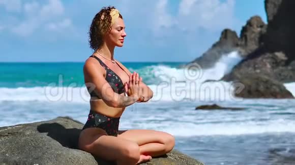 美丽的金发女孩穿着黑色上衣在瑜伽中放松在紫色的垫子上摆莲花对抗海浪在岩石上奔跑视频的预览图