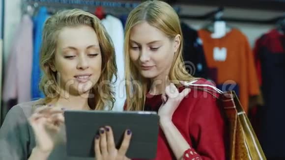 两个女顾客在商店里享受平板电脑站在背景站着穿着衣服手持购物视频的预览图