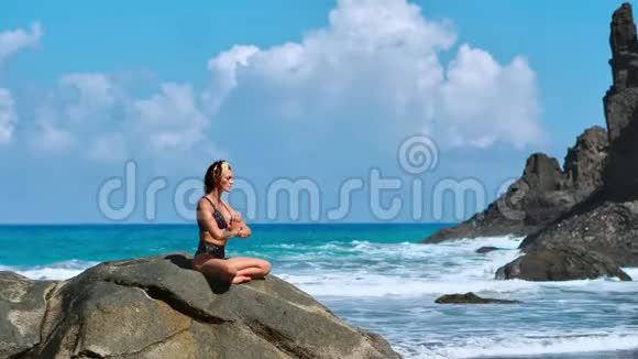 美丽的金发女孩穿着黑色上衣在瑜伽中放松在紫色的垫子上摆莲花对抗海浪在岩石上奔跑视频的预览图