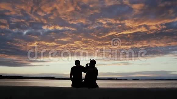 一对年轻情侣坐在海边散步的剪影背景是美丽的日落天空视频的预览图