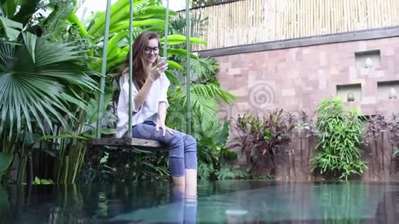 在热带国家的游泳池附近坐在秋千上的女人使用智能手机视频的预览图