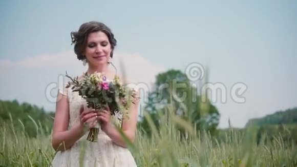 一位美丽的年轻新娘站在麦田里看着她的婚礼花束穿着漂亮的蕾丝婚纱视频的预览图