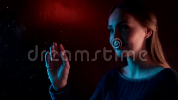 忧郁的年轻的梦幻女子靠近蒸汽玻璃窗在蓝色的霓虹灯中吸引着心灵在她身后的女孩视频的预览图