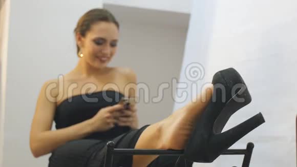 穿着黑色晚礼服的商务女性放松地坐着用智能手机晃着一只高跟鞋微笑着视频的预览图