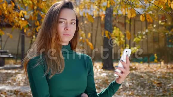秋天一位年轻漂亮的黑发女人带着智能手机在黄树背景下的肖像视频的预览图