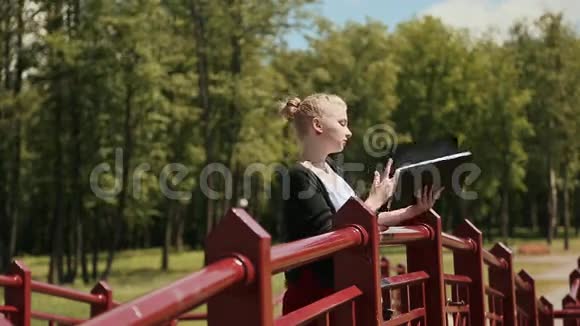 年轻漂亮的女孩在桥上的绿色公园里看书金发视频的预览图