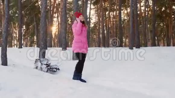 穿着粉红色羽绒服的年轻女子在冬天的树林里背着一个空雪橇用手机说话视频的预览图