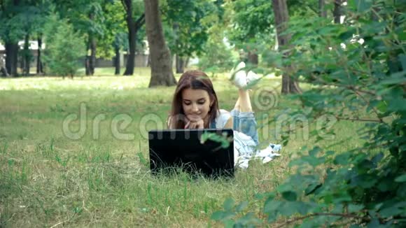 美丽的女士躺在公园的草地上看着笔记本电脑视频的预览图