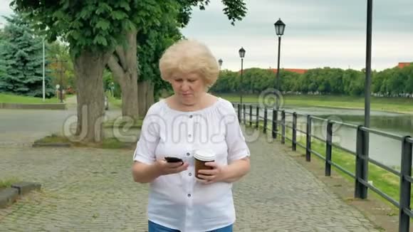 快乐的老妇人使用智能手机和喝咖啡沿着海滨散步视频的预览图