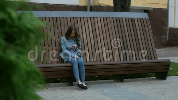 一个严肃的女孩坐在长凳上穿着牛仔裤用手机发短信视频的预览图