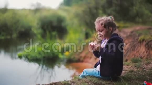 一个可爱的小女孩坐在河岸上吃西瓜背景中的模糊自然侧视图视频的预览图