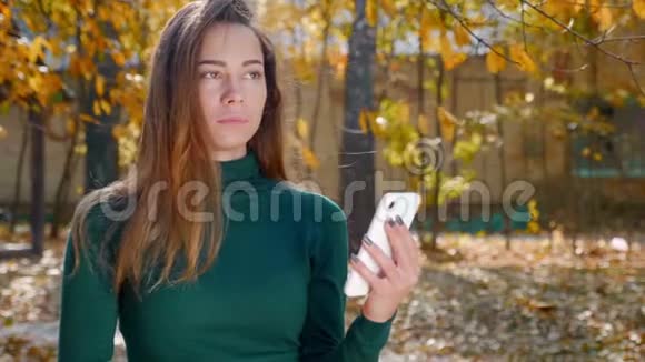 秋天一位年轻漂亮的黑发女人带着智能手机在黄树背景下的肖像视频的预览图