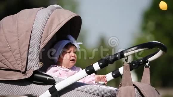 在阳光明媚的日子里哭泣的女婴坐在公园里的婴儿车厢里视频的预览图