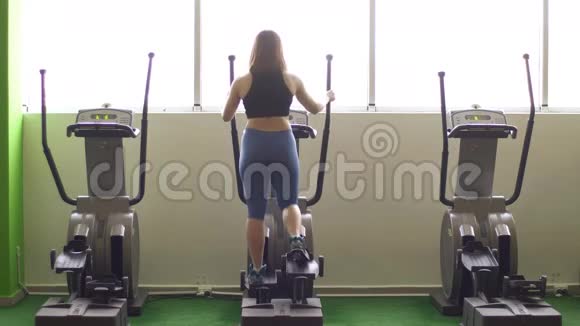 这位女士在健身房窗户后面的椭圆训练器上工作的后视图视频的预览图