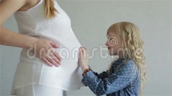 穿着蓝色裙子的小女孩正在抚摸和亲吻她母亲的大肚子视频的预览图