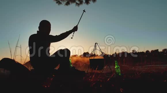 男子旅游舞蹈正坐在篝火晚会的大自然露营剪影阳光日落度假时的男性剪影视频的预览图