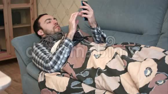 生病的年轻人拿着手机躺在沙发上视频的预览图