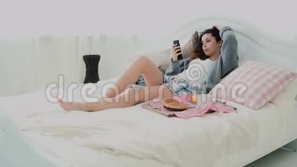 年轻微笑的女人躺在床上布鲁内特女孩在早餐期间上网冲浪使用智能手机和饮料果汁视频的预览图