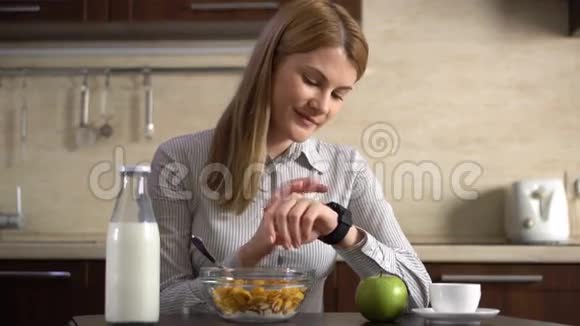 漂亮的女商人吃早餐用她的智能手表浏览互联网在看新闻视频的预览图