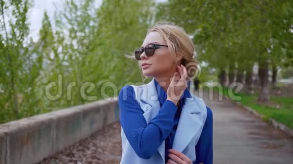 美丽的金发商务女性戴着太阳镜穿着蓝色西装在河边等着在城里见面放松一下视频的预览图